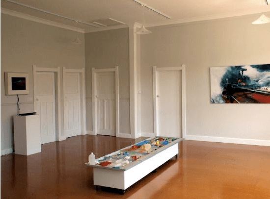 A long table full with objects in a large room with four white doors.